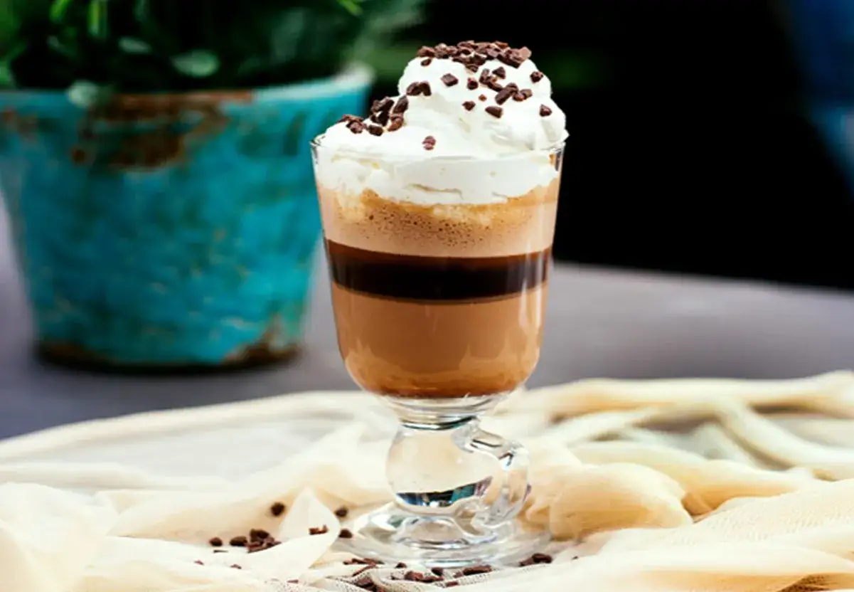 Un bicerin coffee, stratificato e guarnito con panna montata e scaglie di cioccolato, in un bicchiere elegante.