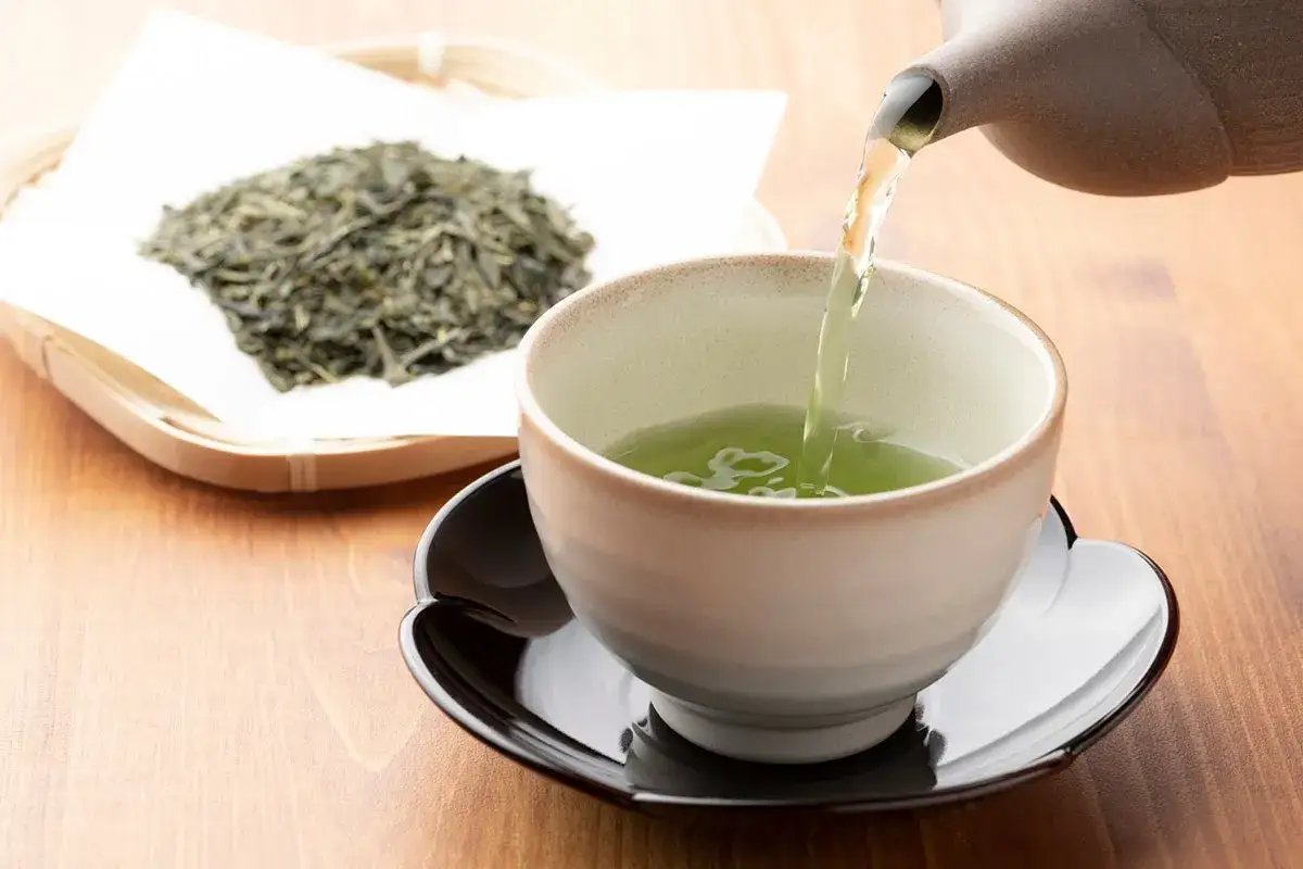 Tè verde versato in una tazza, con foglie secche accanto. Una bevanda salutare che aiuta a combattere il colesterolo.