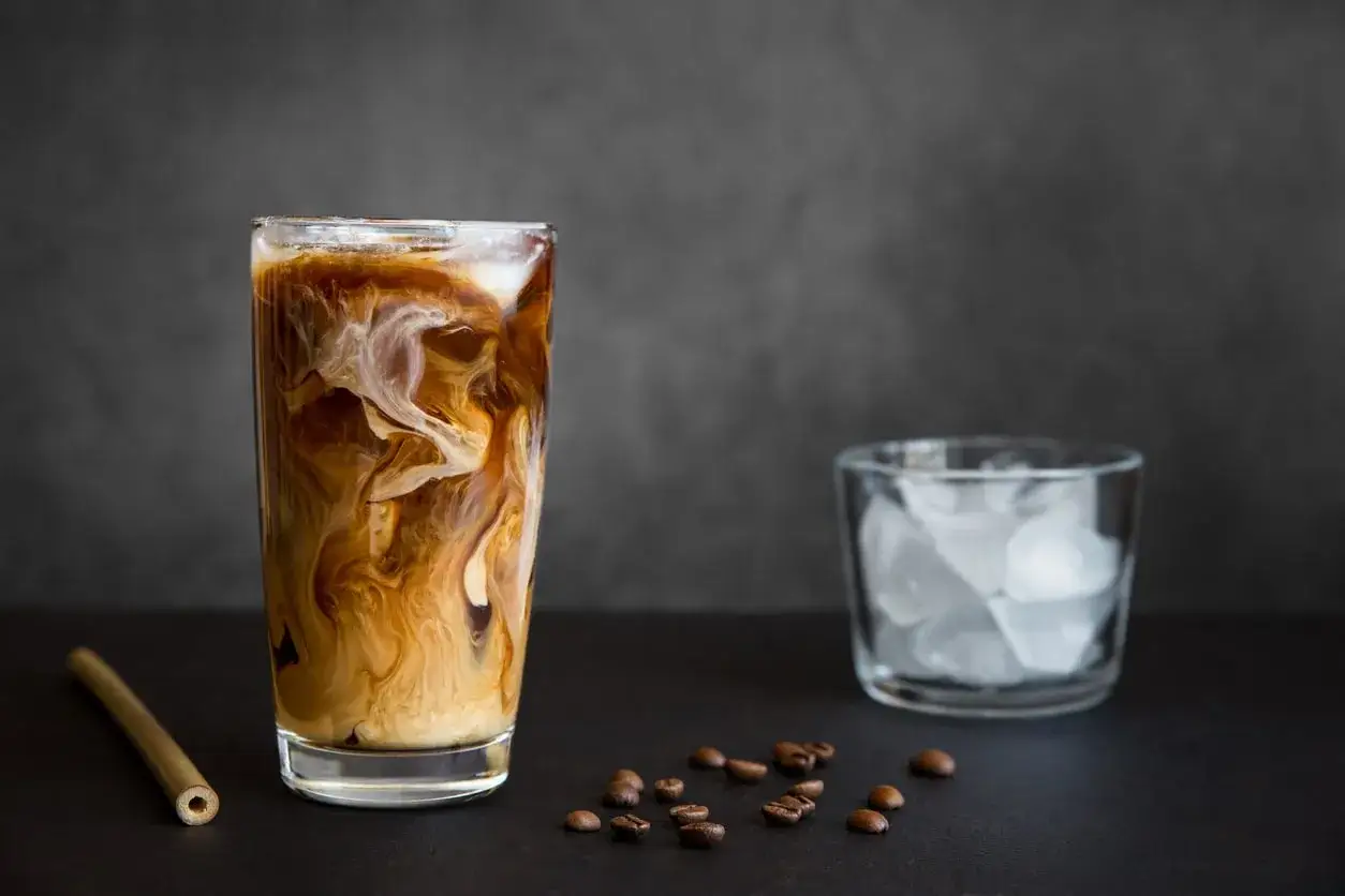 Iced coffee come si fa: un bicchiere di caffè freddo con latte e ghiaccio, chicchi di caffè e un bicchiere di ghiaccio.