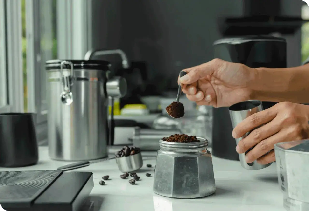 Mano aggiunge caffè macinato a una moka, per il suo primo utilizzo. Caffè in grani e contenitori in acciaio completano la scena.