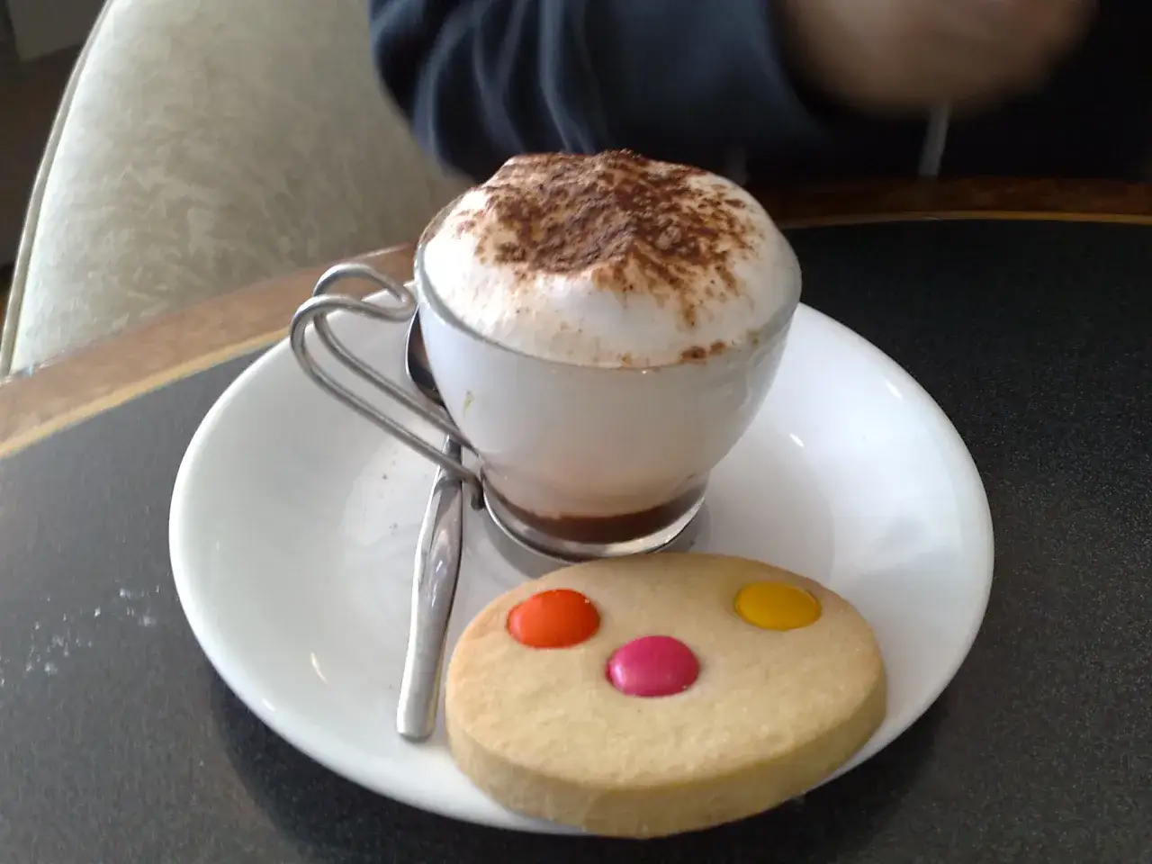Un delizioso cappuccino al bar, con schiuma abbondante e cacao, accompagnato da un biscotto decorato con caramelle colorate.