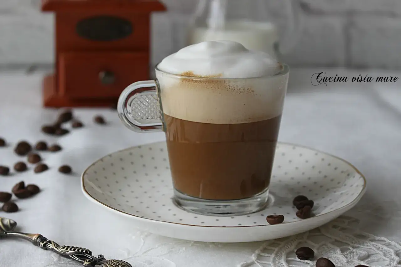 Un bicchiere di caffè macchiato vs cappuccino, con schiuma soffice, chicchi di caffè sparsi e un macina caffè in legno sullo sfondo.