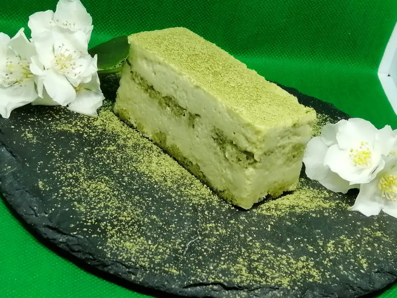 Un delizioso matcha tiramisù, cosparso di polvere verde, adagiato su una pietra scura con fiori bianchi.