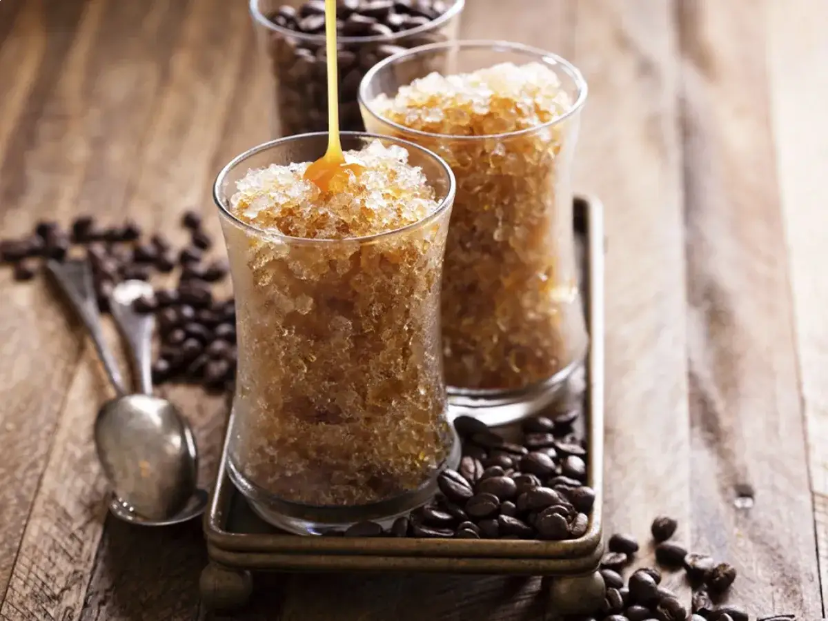 Granita di caffè fatta in casa, con sciroppo dorato che cola su una delle coppe. Chicchi di caffè tostati e cucchiaini completano la scena.