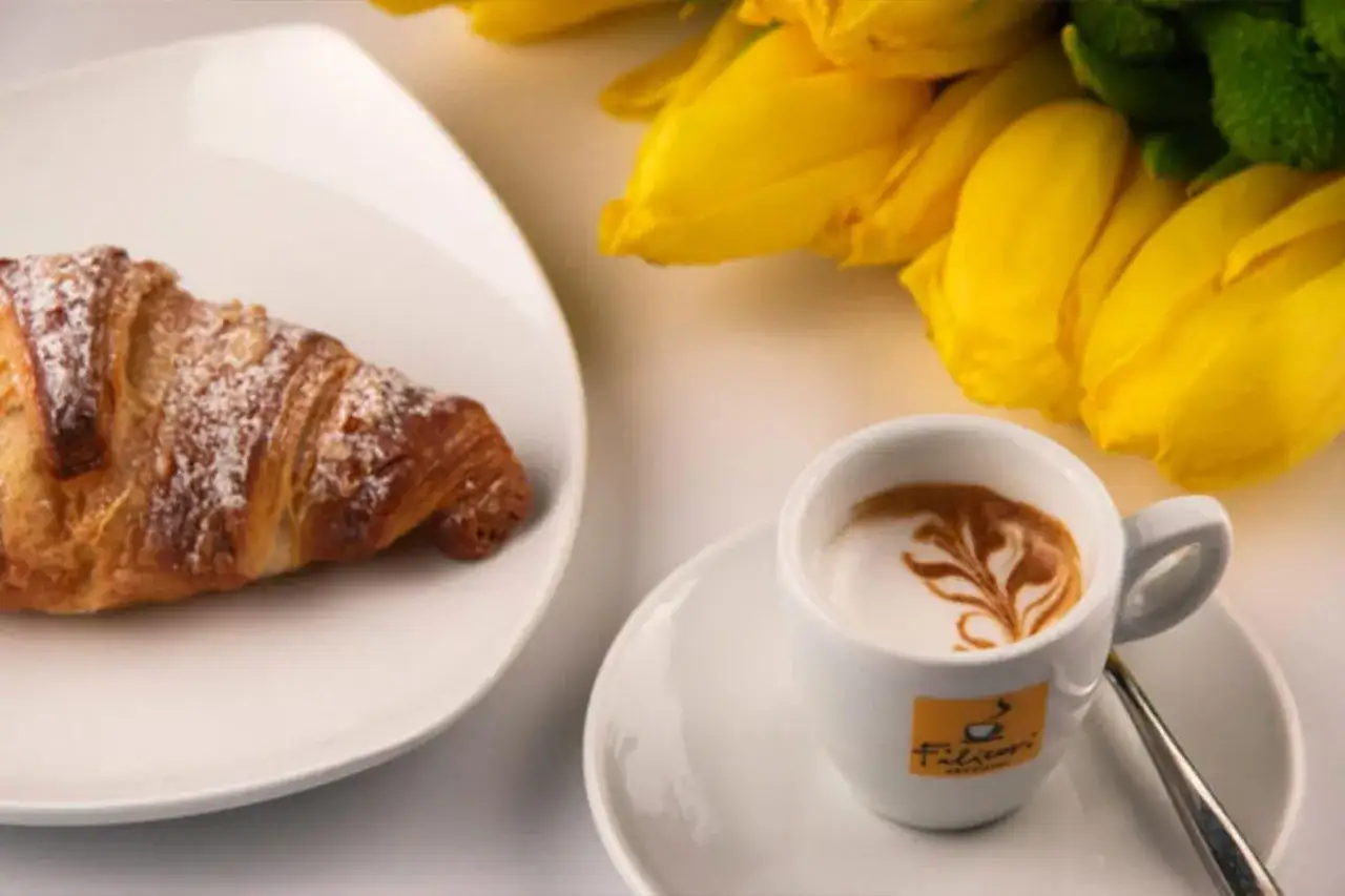 Un croissant spolverato di zucchero a velo e un caffè macchiato con schiuma decorata, accanto a tulipani gialli.
