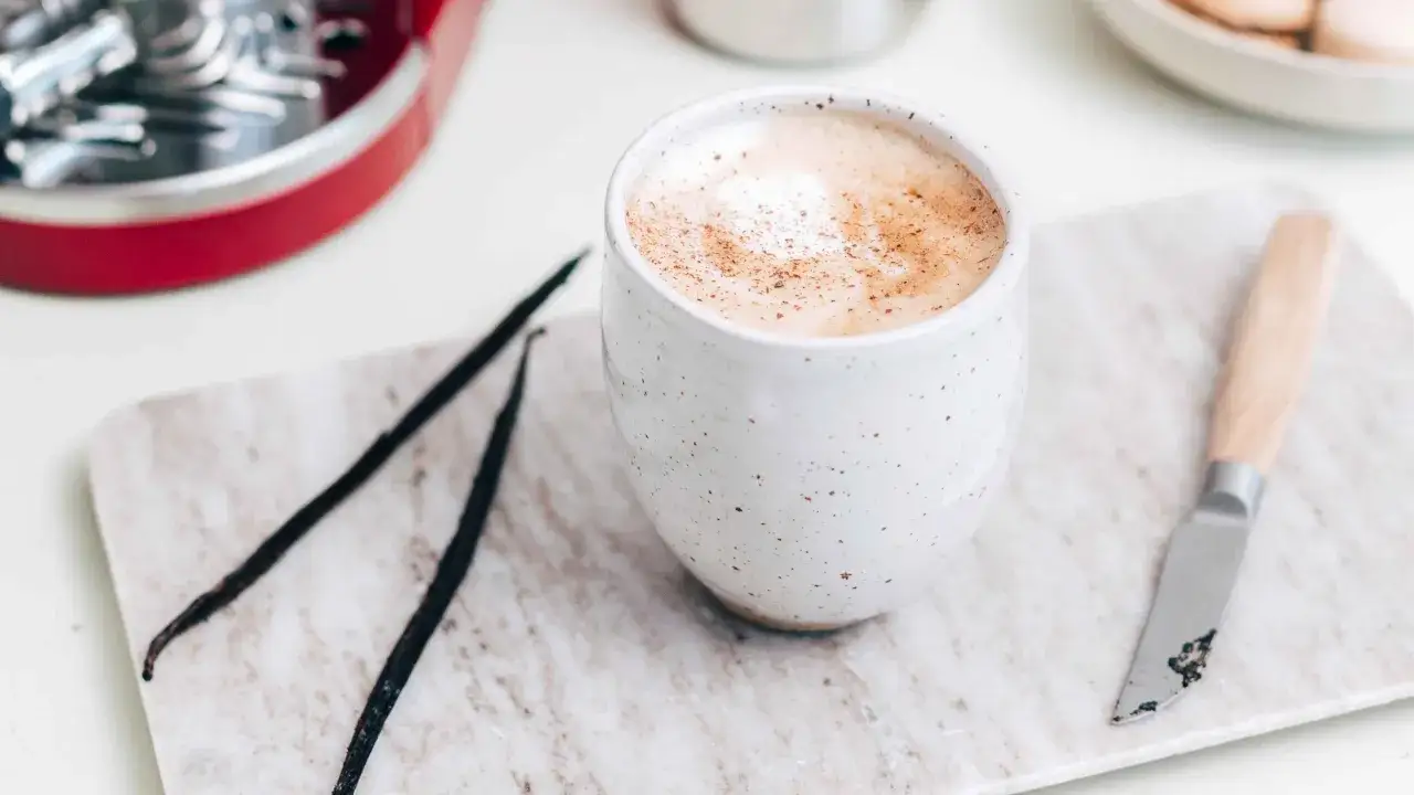 Un cremoso cafe latte spolverato di cannella, accanto a baccelli di vaniglia e un piccolo coltello su un tagliere di marmo.