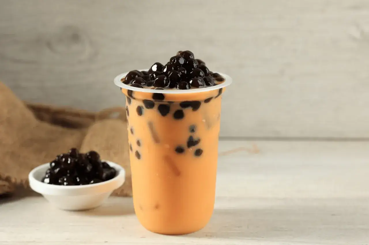 Un bicchiere di bubble tea arancione con perle nere, un piccolo contenitore di perle e un telo di juta. La sua origine &egrave; un viaggio di sapori.