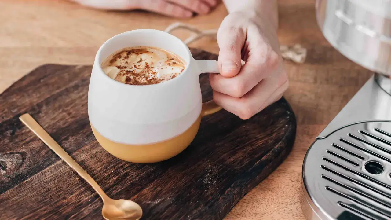 Mug con schiuma e cannella, un cucchiaino dorato e una macchina per caffè. La ricetta del pumpkin spice latte Starbucks.