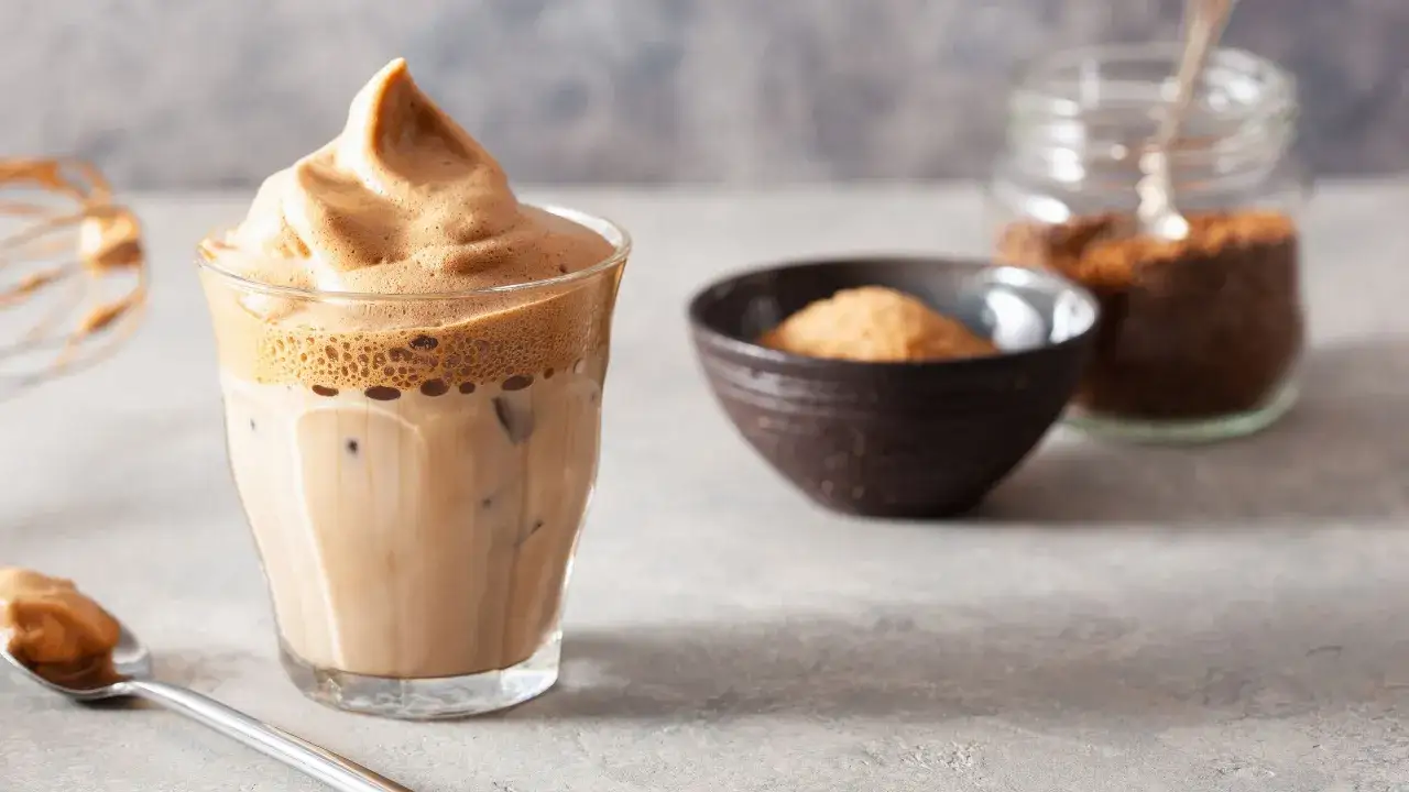 Un bicchiere di caff&egrave; freddo con una generosa spuma di caff&egrave; in cima. Accanto, una ciotola con zucchero e un barattolo di caff&egrave; solubile.