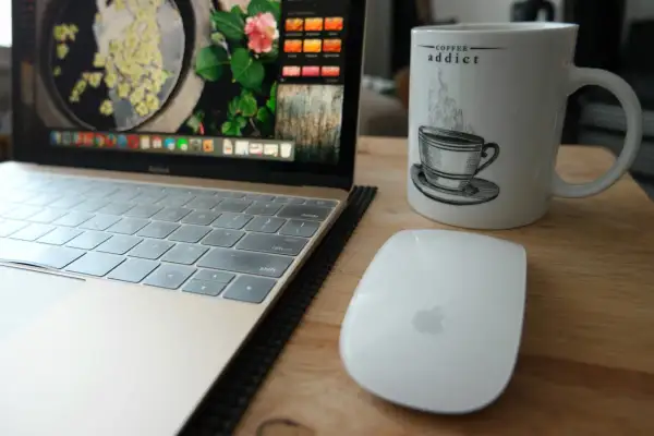 Tazza con scritta "Coffee addict", laptop e mouse su un tavolo di legno. La dipendenza da caffeina è evidente.