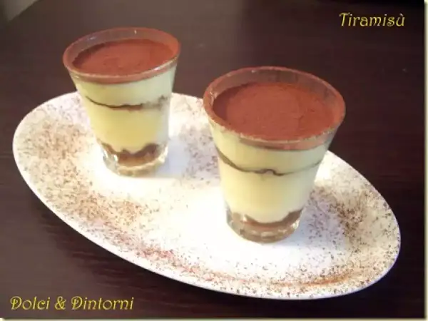 Due tiramisù monoporzione con savoiardi, presentati in bicchieri e spolverati di cacao, su un piatto bianco.