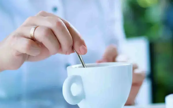 Mano con anello mescola il caffè in una tazza bianca. La scelta è sempre la stessa: caffè con o senza zucchero.