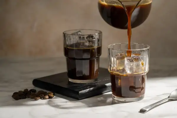Versando caffè freddi in bicchieri con ghiaccio. Chicchi di caffè tostati e un cucchiaino completano la scena.