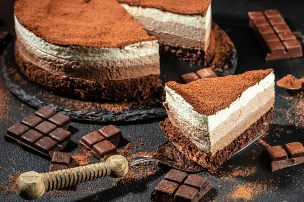 Una golosa cheesecake cappuccino a tre strati, cosparsa di cacao, con pezzi di cioccolato e una fetta servita su una forchetta.