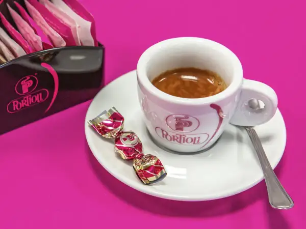 Una tazzina di crema al caffè espresso, con tre caramelle incartate e un porta-bustine Portioli su sfondo fucsia.