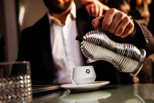 Un uomo in giacca e camicia versa caffè da un bricco argentato in una tazzina bianca. La macinatura caffè più fine garantisce un aroma intenso.
