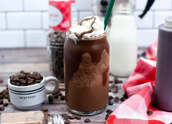 Un delizioso Frappuccino Java Chip, con panna montata e sciroppo al cioccolato. Sullo sfondo, chicchi di caffè, gocce di cioccolato e latte, tutti ingredienti per prepararlo.