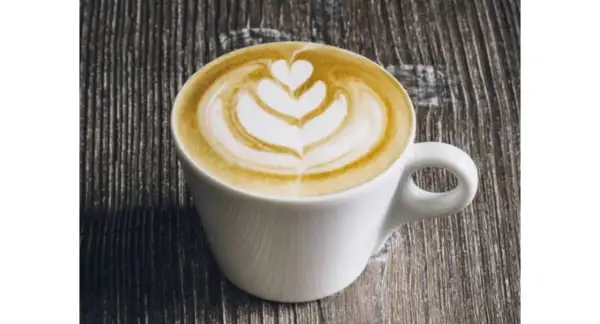 Un flat latte con schiuma artistica a forma di cuore e petali, servito in una tazza bianca su un tavolo di legno scuro.