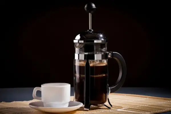 Una tazzina bianca accanto a una caffettiera francese piena di caffè scuro, pronta per essere gustata.