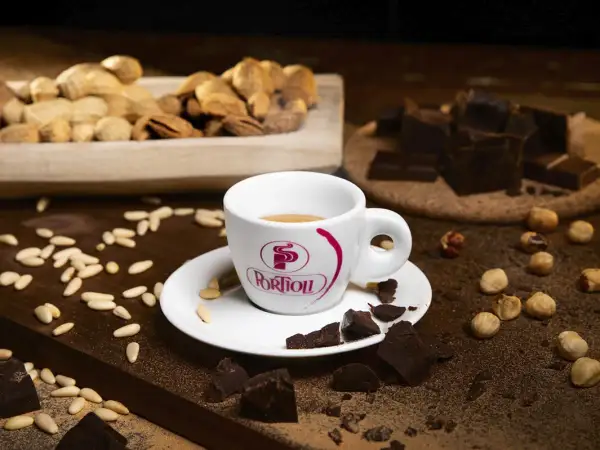 Tazza di caffè fumante, circondata da pezzi di cioccolato fondente, nocciole e pinoli. Un'invitante combinazione di caffè e cioccolato.