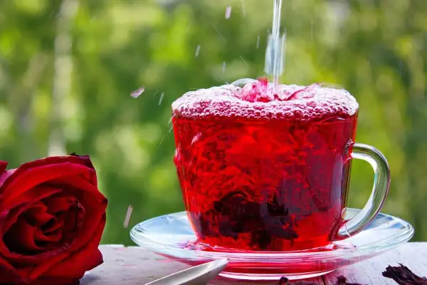 Tè rosso vibrante versato in una tazza, con una rosa rossa accanto. Scopri le proprietà e le controindicazioni dell'ibisco.