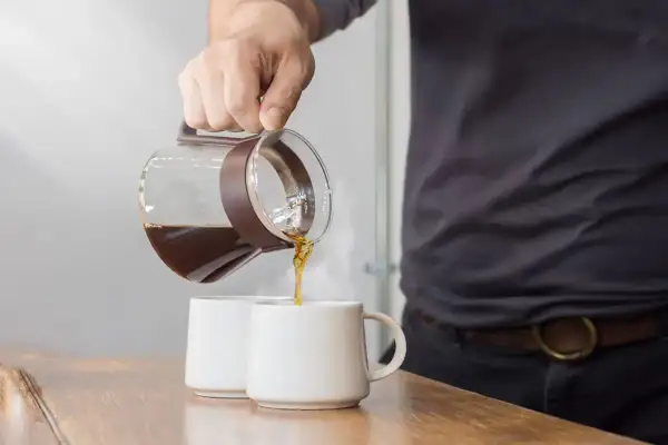 Un uomo versa caffè americano con latte in due tazze bianche su un tavolo di legno.