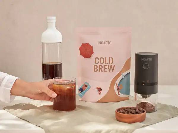 Un bicchiere di caffè freddo in bottiglia, pronto per essere gustato. Accanto, una confezione di cold brew e un macinacaffè.