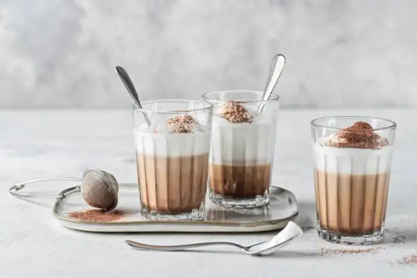 Tre bicchieri di cappuccino con cioccolato, guarniti con panna e cacao, su un vassoio con colino.